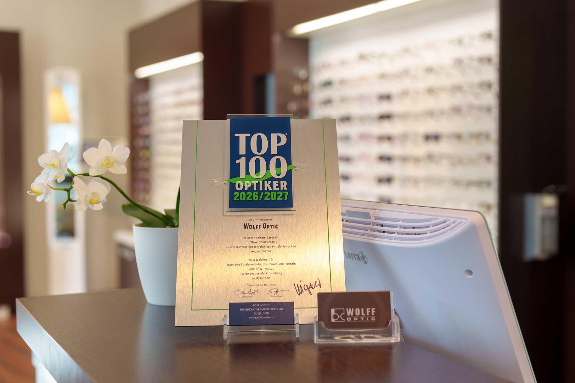 Wolff-Optic-0326_Top100-Urkunde Aufsteller „Top 100 Optiker 2024/2025“ steht auf Theke, daneben Blumentopf und Firmenkarte.